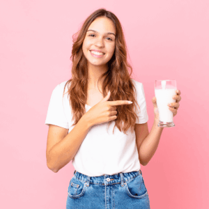Eine junge Frau mit Laktoseintoleranz zeigt freudestrahlend auf ein Glas frischer Milch in ihrer Hand.